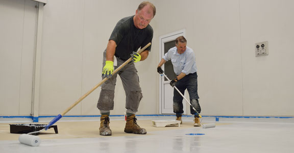 This image shows 2 men painting a floor with a roller brush.
